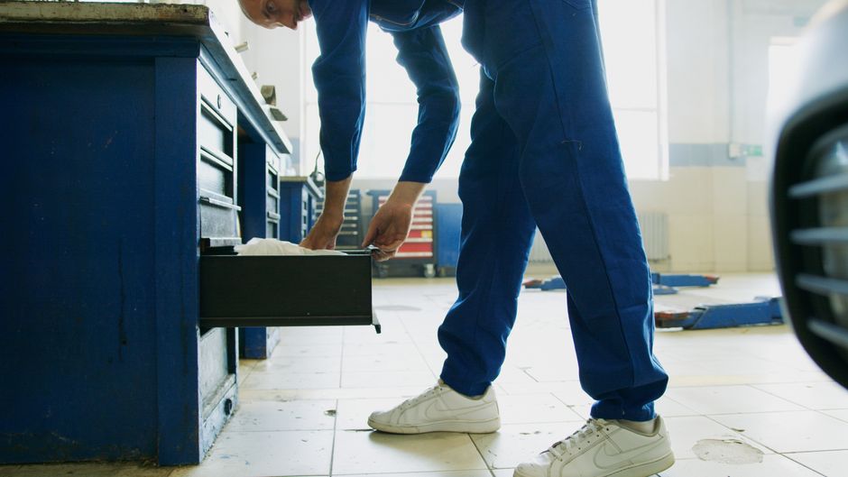 Mechanic in blue coveralls opens a toolbox drawer in a sunlit workshop.